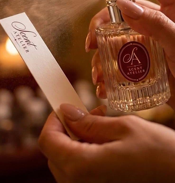 Parfem se rasprskava na mirisnu trakicu uz svjetlo svijeća, s bočicom Scent Atelier u drugoj ruci.